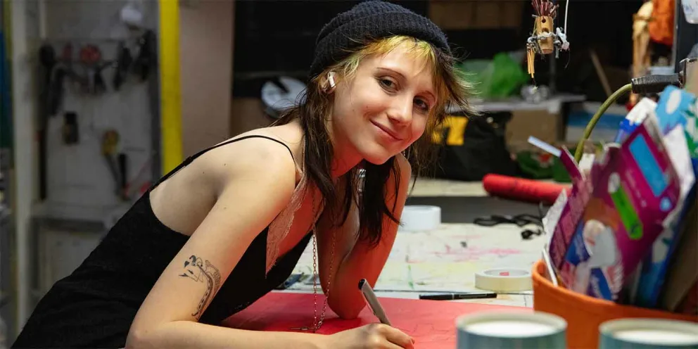 Person in a black dress and beanie, leaning over a table, writing on red paper, surrounded by art supplies and craft materials.
