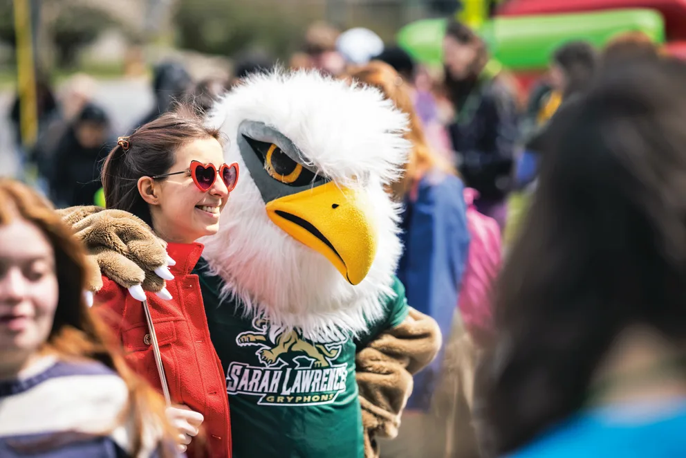 Person with heart-shaped sunglasses posing with an eagle mascot wearing a Sarah Lawrence Gryphons shirt at an outdoor event.