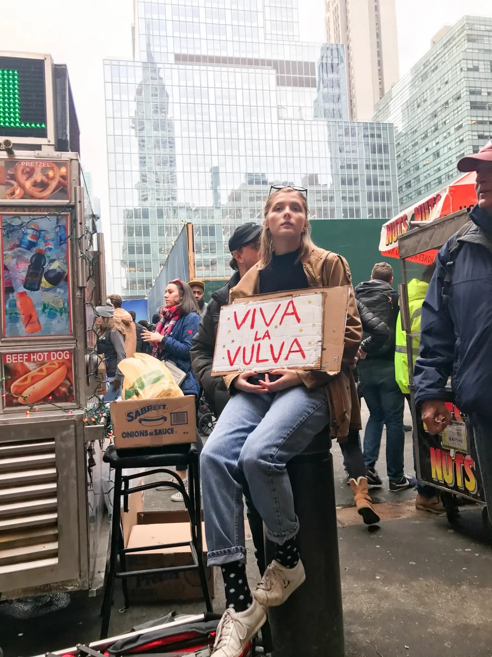 Person sitting on a bollard holding a sign that says "Viva La Vulva" near food carts in an urban setting.