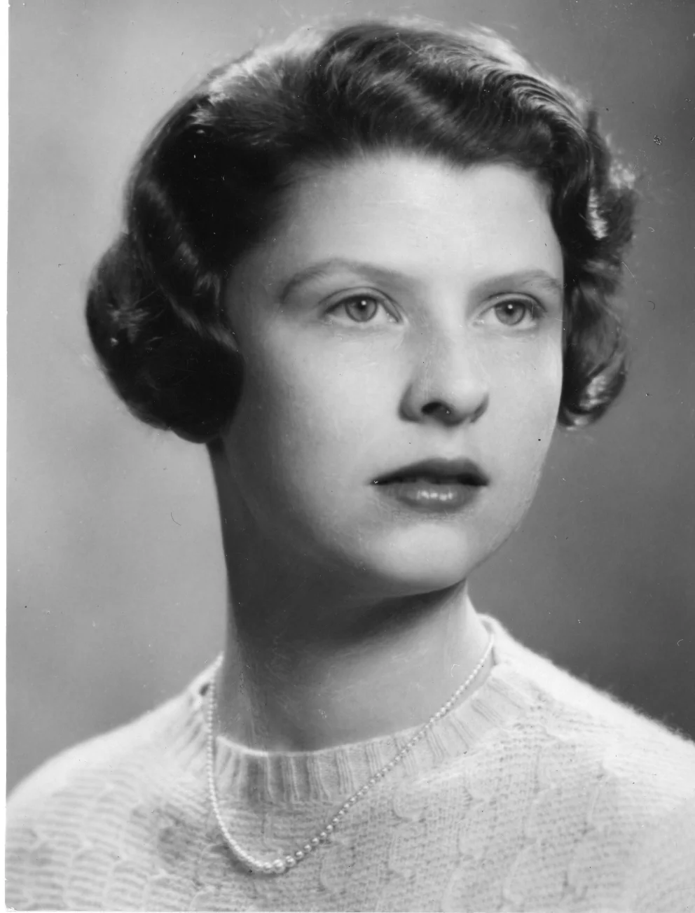 Individual with short, wavy hair, wearing a light sweater and pearl necklace, gazes slightly to the side in a black and white portrait.