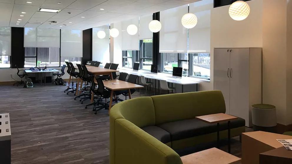 Modern office space with green couches, desks with computers, and white globe lights. Large windows with blinds.