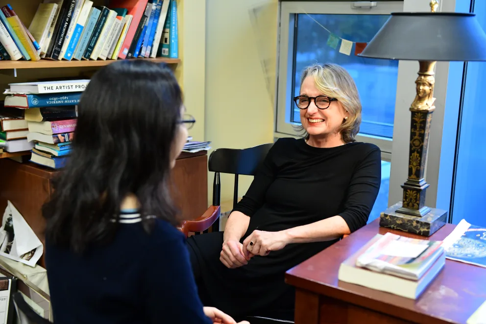 Two people sit in a cozy room filled with books. One faces away, the other smiles and relaxes in a chair by a desk with a lamp.