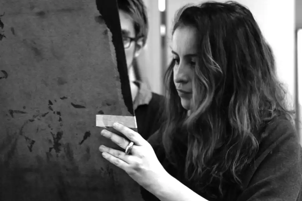 Person examining a large sheet of paper with focus, another person partially visible in the background. Black and white image.