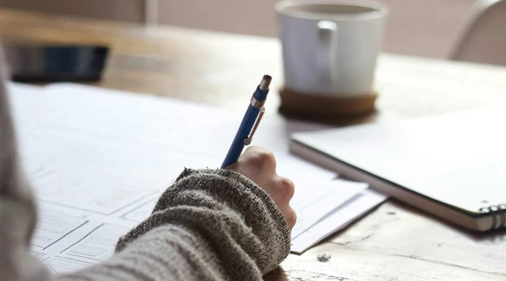 Person writing on paper at a wooden table with a pen, wearing a sweater. A cup and notebook are on the table in the background.