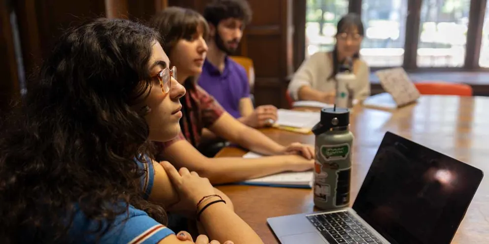 Four people sitting at a table, engaged in discussion, with a laptop and water bottle visible in the foreground.