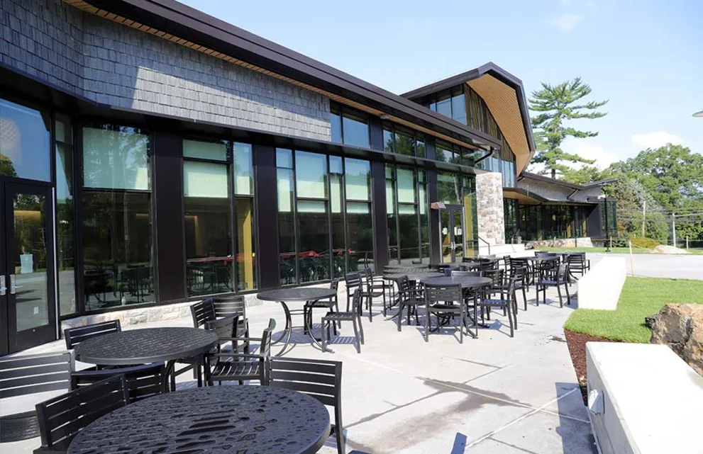 Outdoor seating area with round tables and chairs in front of a modern building with large glass windows and a stone exterior. Trees in the background.