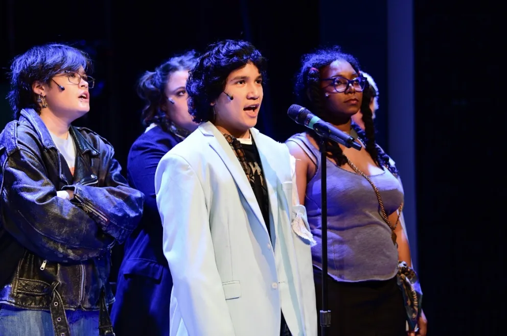 Three performers on stage, one in a blue suit singing into a microphone, flanked by others in casual attire under bright stage lights.