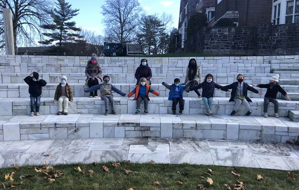 People sitting spaced apart on stone steps outdoors, wearing masks and holding their arms out.