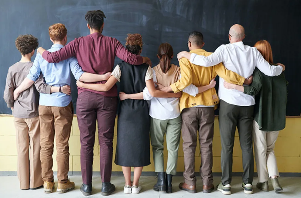 Eight people standing in a line, arms around each other, facing a chalkboard.