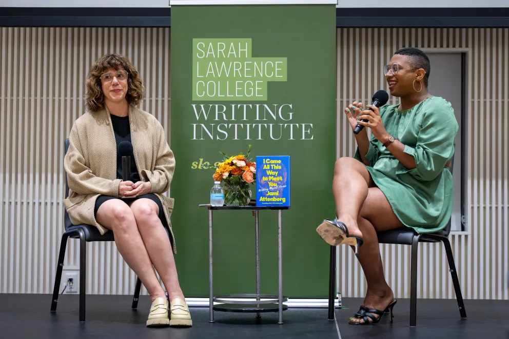 two women sitting on chairs talking to each other in front of a green wall with a sign behind them