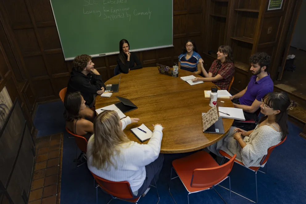 A group of people sits around a circular wooden table in a meeting room with a green chalkboard, taking notes and using laptops.
