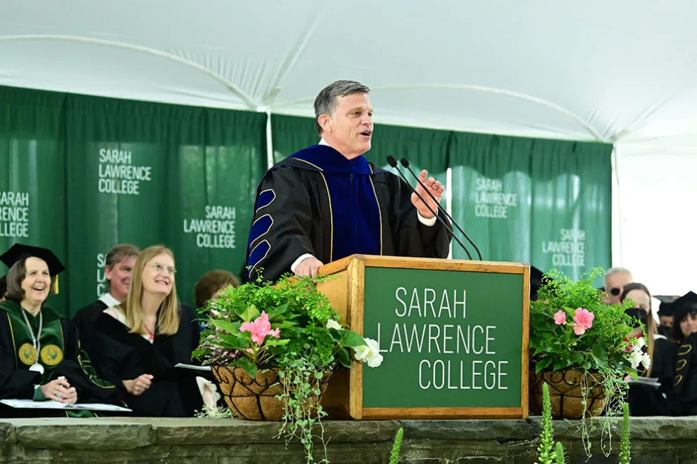 Speaker at a podium with Sarah Lawrence College signage, addressing a seated audience in academic regalia under a tent with green banners.