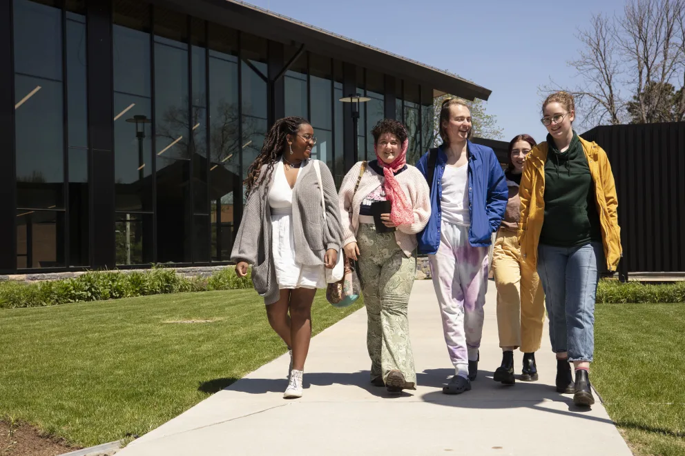 Five people walking and smiling on a sunny day, outside a modern building with large glass windows and surrounded by greenery.