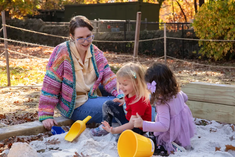 Person and two children play with buckets in a sandbox on a sunny autumn day, surrounded by colorful leaves.