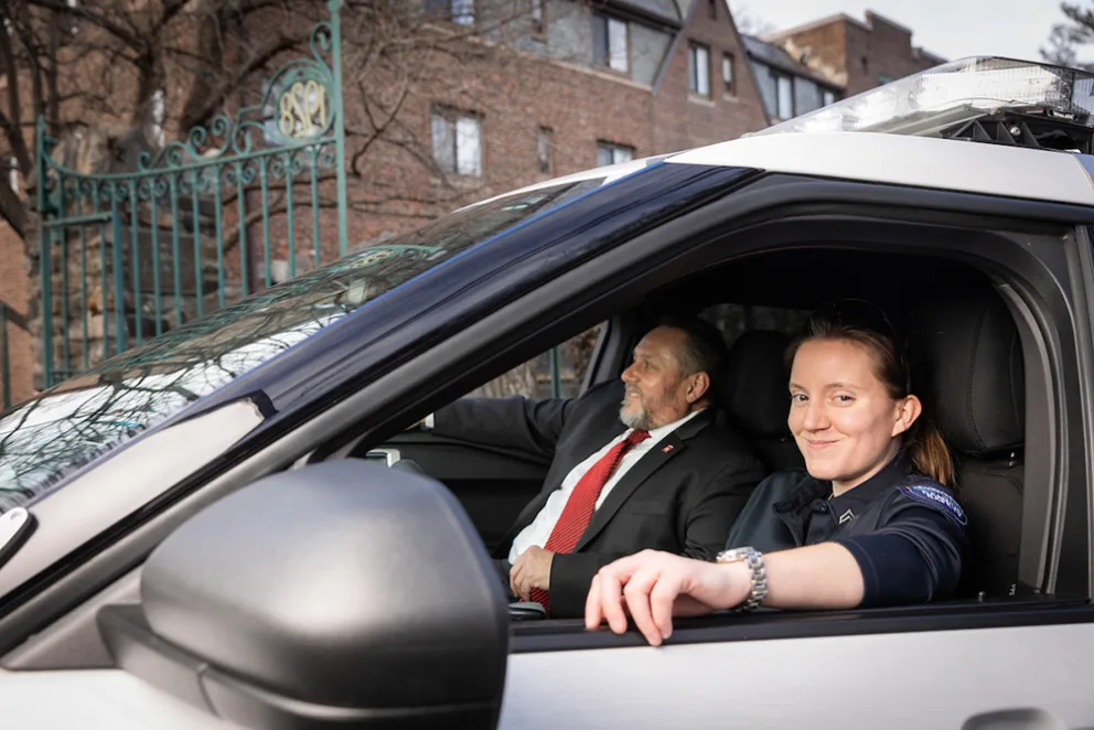 Two people sitting in a parked police vehicle. The driver looks toward the camera, while the passenger looks out the window. Urban setting in the background.