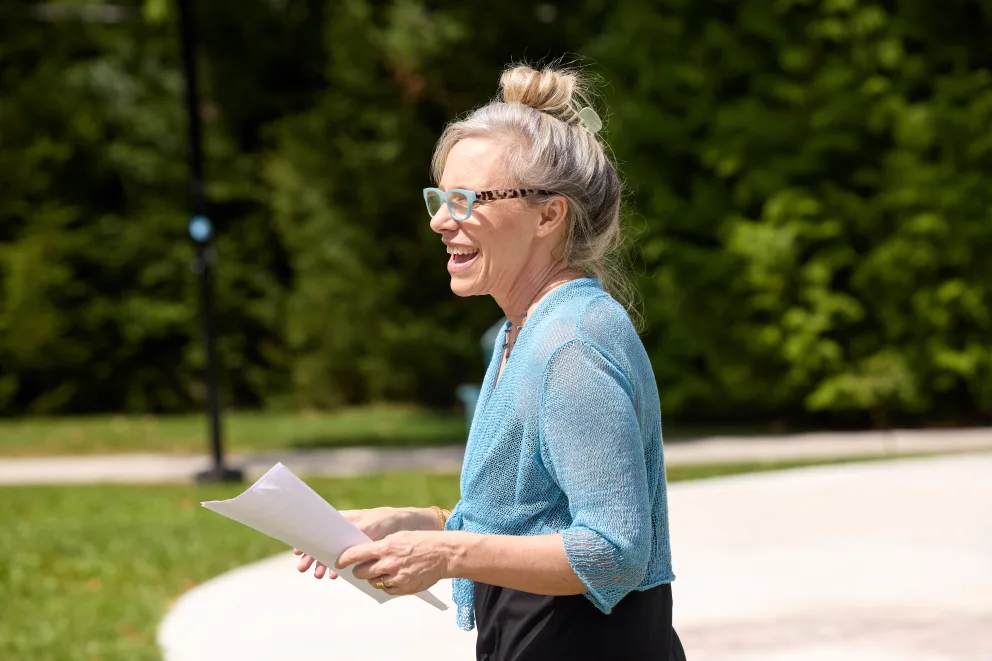 Person outdoors in a park, wearing glasses and a blue top, holding papers, and smiling, with trees in the background.