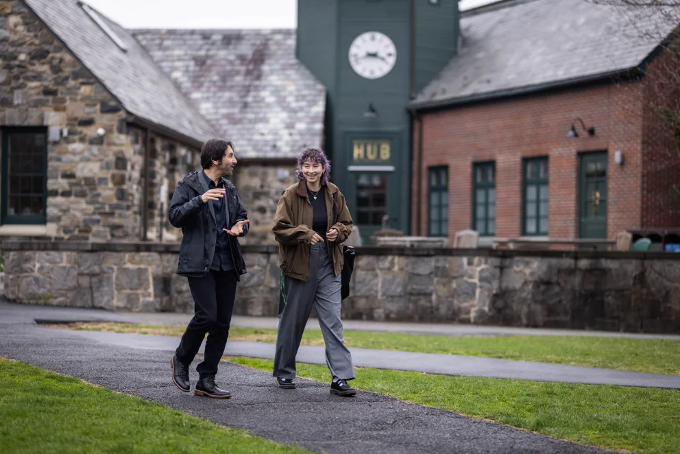 Two people walk and talk on a path in front of stone buildings with a clock tower. One wears a dark jacket; the other, a brown jacket.