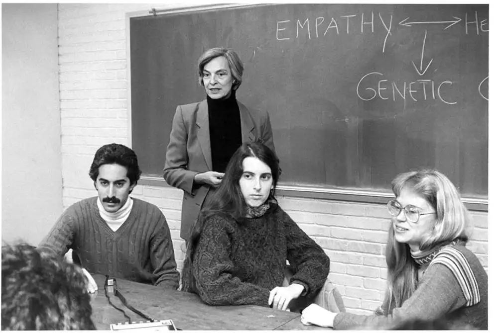 In a classroom, a person stands near a blackboard with "Empathy" written on it, while three others sit at a table facing forward.