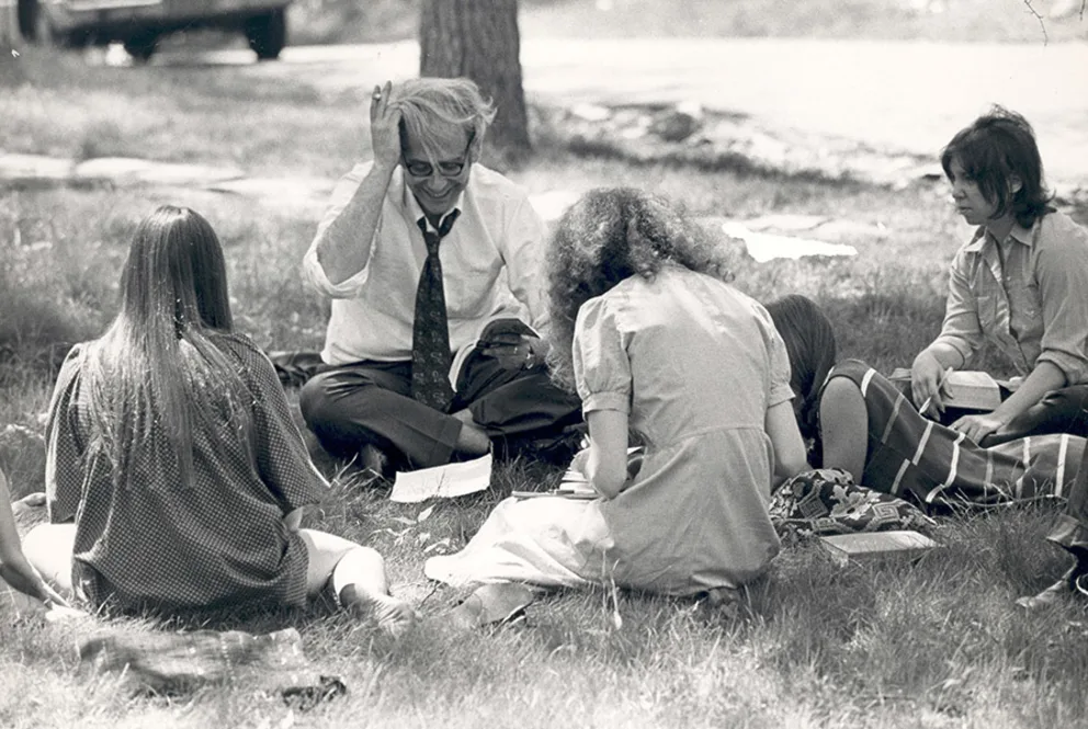 Group of people sitting on grass with books, engaged in discussion. A person in the center adjusts their glasses. Trees and parked car in background.