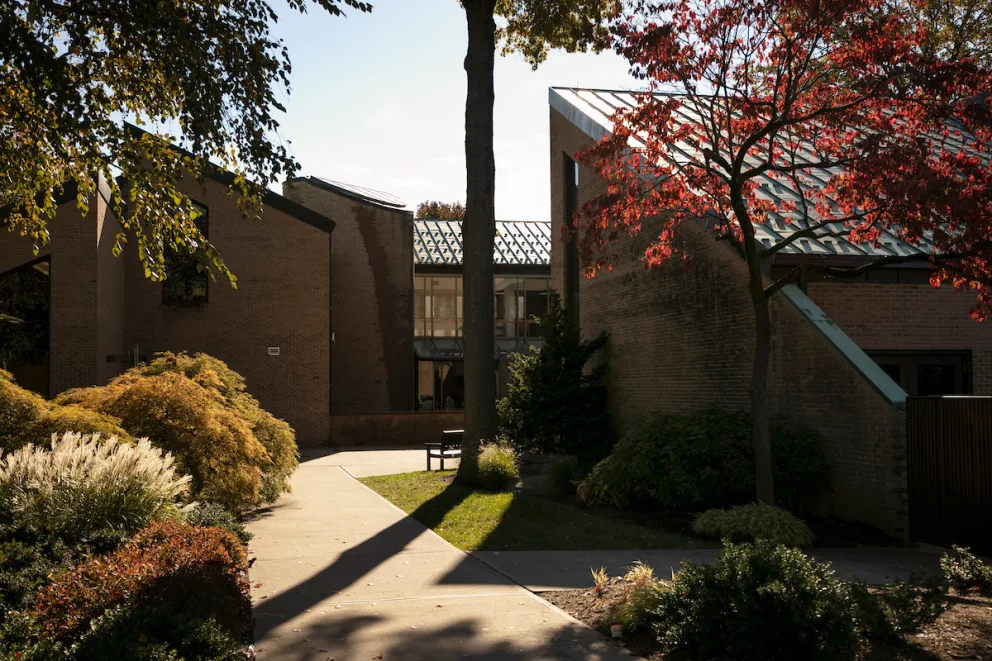 Pathway between modern brick buildings with greenery and trees, including one with red leaves, under a clear sky.
