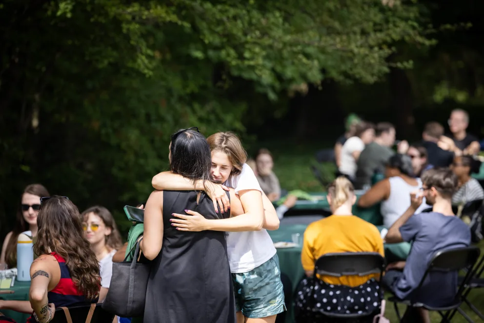 People gather outdoors. Two individuals hug in the foreground. Others sit at tables, enjoying the sunny weather and greenery.