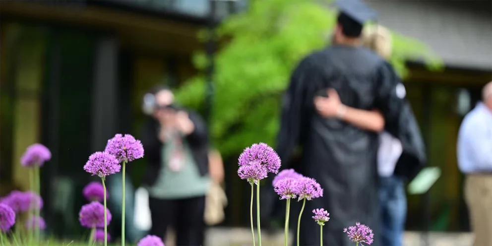 Purple flowers in focus with blurred background of people; one in cap and gown, embracing another in a photo pose.