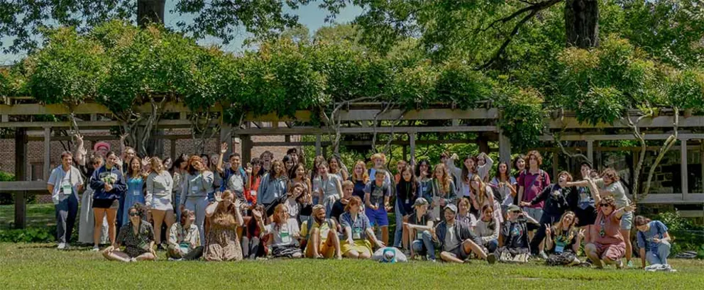 A large group of people posing together on a grassy area with trees and a wooden structure in the background.