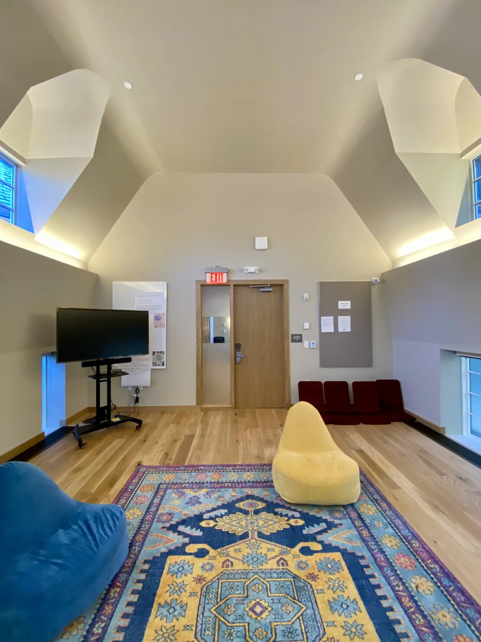 Room with high ceiling, wooden floor, colorful rug, blue and yellow bean bags, mounted TV, and small red chairs against the wall.