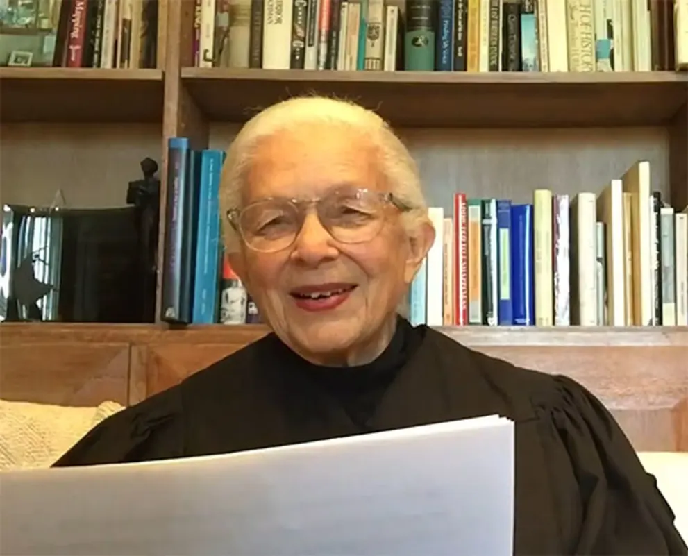 Smiling person holding papers, wearing glasses and a black robe, with a background of a bookshelf filled with various books.