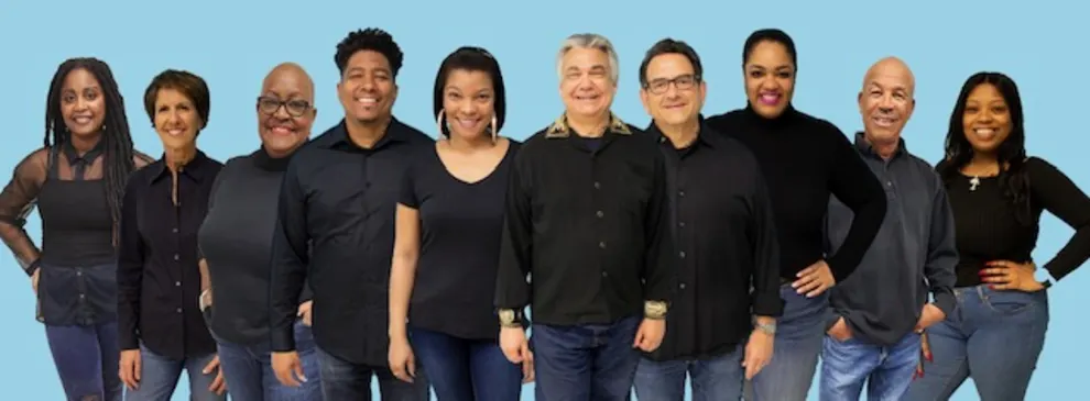 Ten people standing in a line, smiling in front of a blue background, all wearing black tops and jeans.