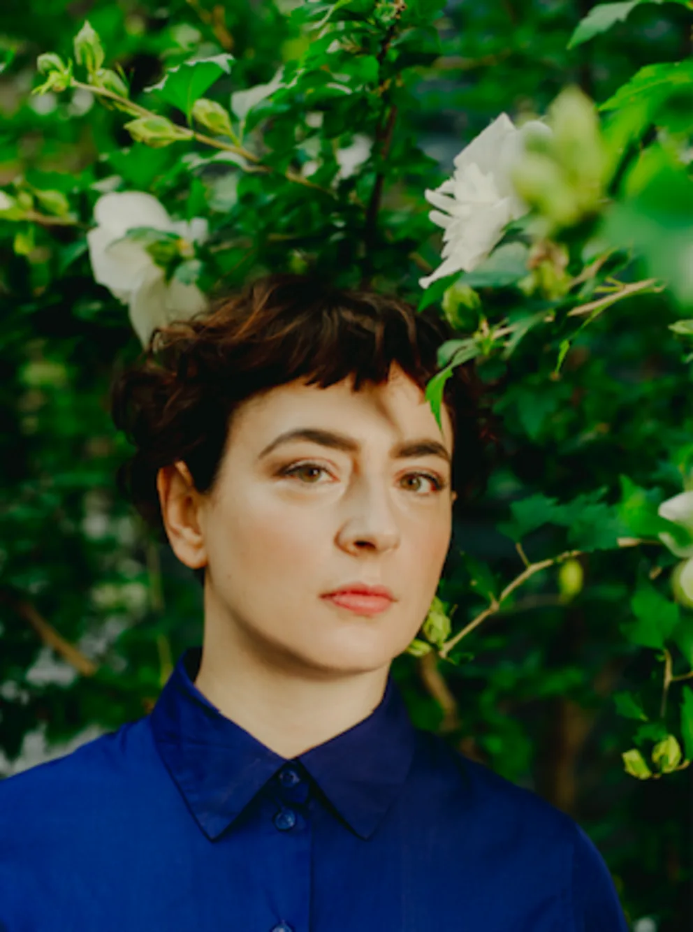 Person in blue shirt stands among green foliage with white flowers in the background.