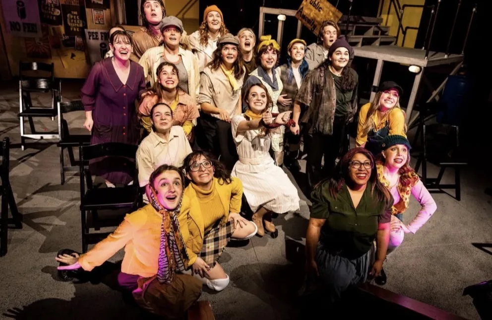Group of performers in costumes, posing energetically on stage with smiles, some pointing or cheering. Black chairs and stage props are visible in the background.
