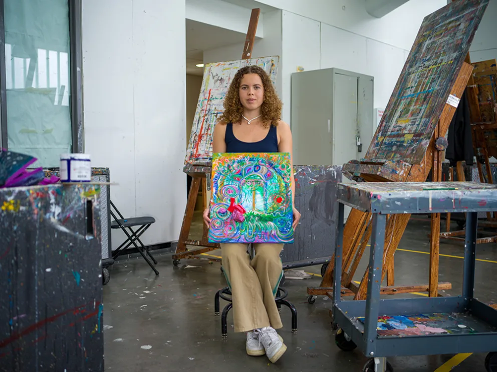 Artist seated in a studio holding a colorful painting, surrounded by easels and art supplies.