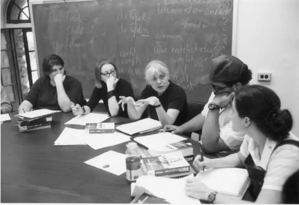 A group of people sit around a table with books and papers, having a discussion, with a blackboard full of notes in the background.