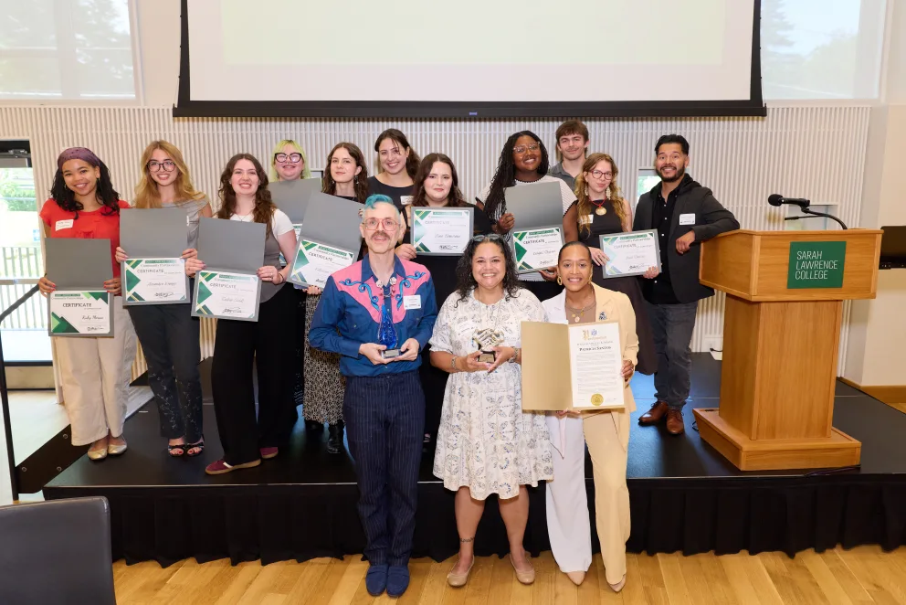 A group of people on a stage holding certificates and awards, standing near a podium with a sign for Sarah Lawrence College.