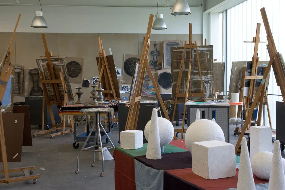 Art studio with easels, canvas paintings, and geometric plaster sculptures on tables under overhead lights.