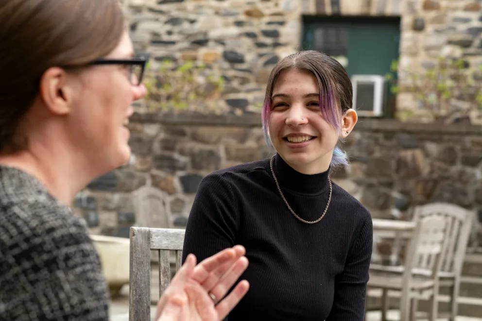 Two people are sitting outside having a conversation. The person in the foreground is gesturing; the other is smiling, wearing a black top.