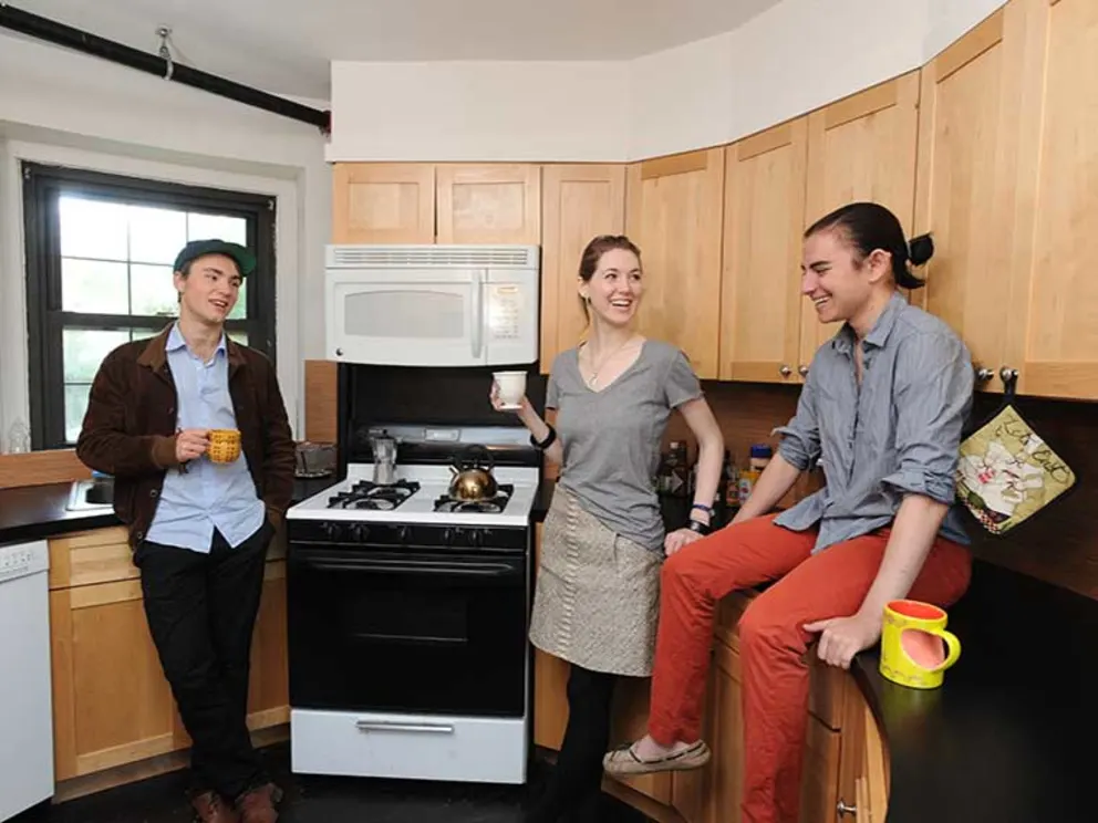Three people in a kitchen talking and smiling, holding mugs. A person sits on the counter, another leans against a stove. Cups are nearby.