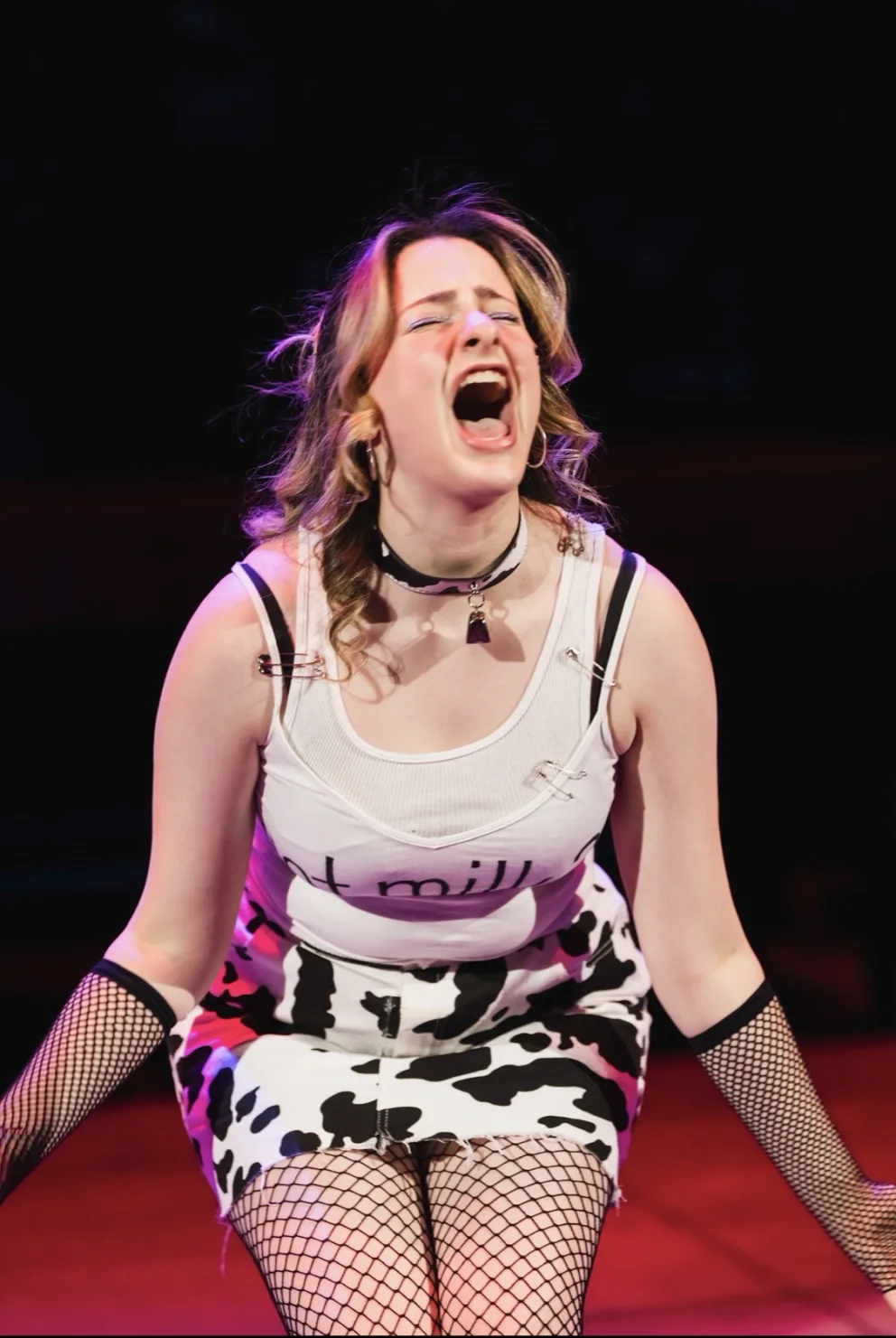 Person on stage with open mouth, wearing a white tank top, cow-print skirt, and fishnet gloves, expressing emotion.