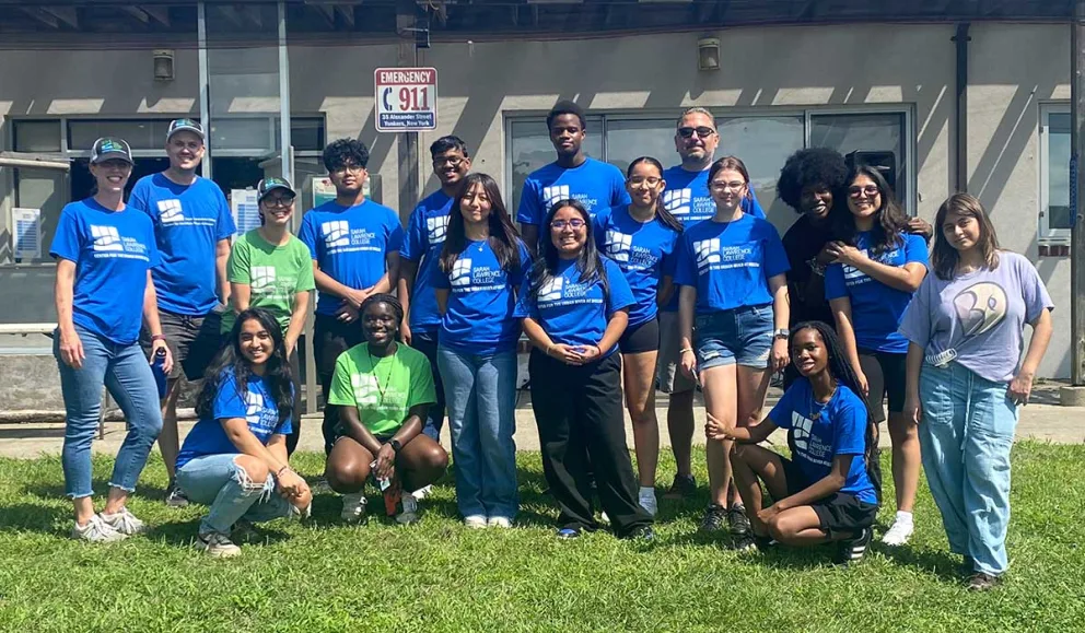 A group of people smiling and posing outdoors, most wearing blue shirts, with one in a light green shirt, on a grassy area in front of a building.