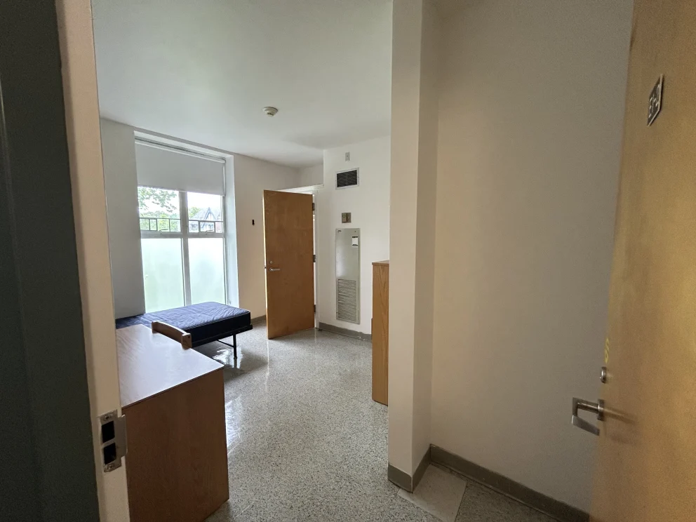Dorm room with an open door, bed, desk, and window. Room is empty, with plain walls and floor, lit by natural light.