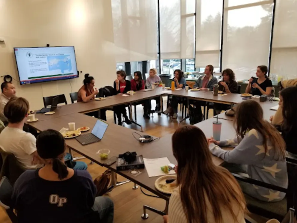 A group of people are sitting around a long table in a classroom, with a laptop on the table and a projector screen in the background.
