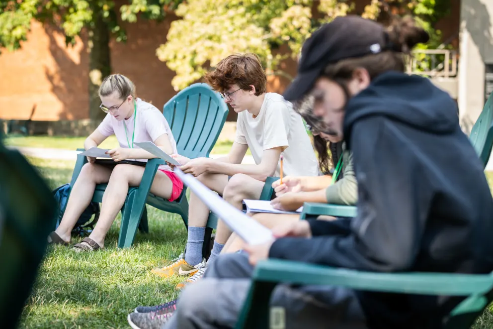 People seated outdoors on lawn chairs, writing on papers, with trees and a building in the background.
