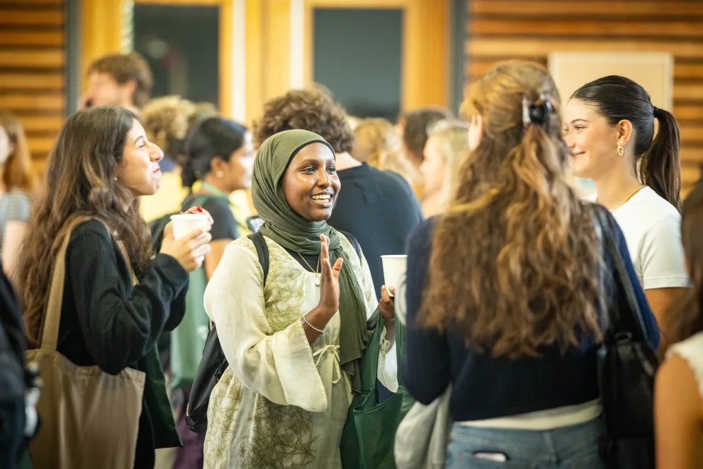 A group of people in conversation at an indoor event, holding drinks and smiling, with a wooden wall background.