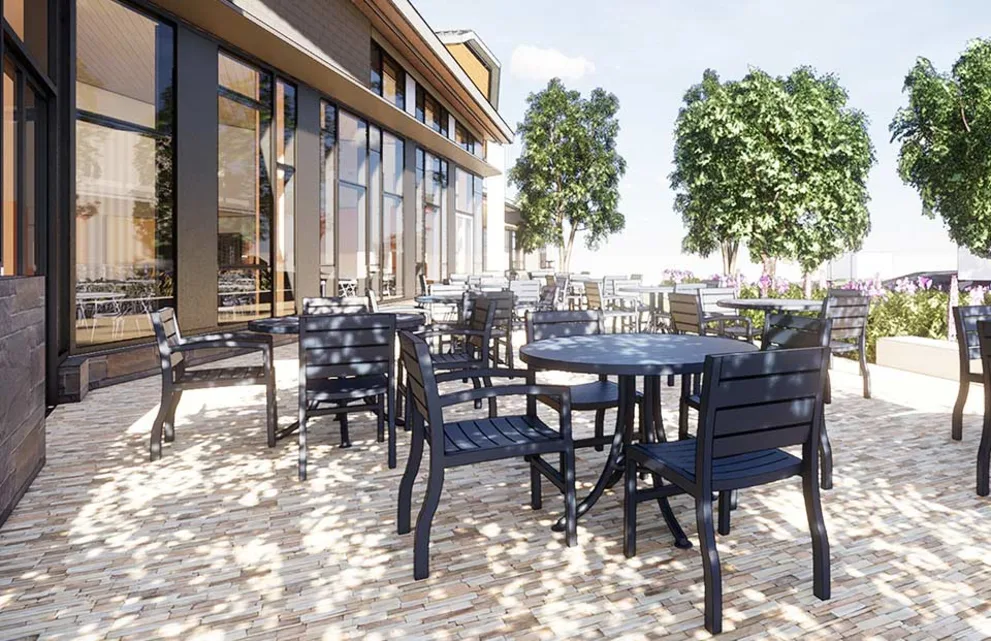 Outdoor patio with black tables and chairs, surrounded by trees. Large windows reflect the clear sky on the building's exterior.