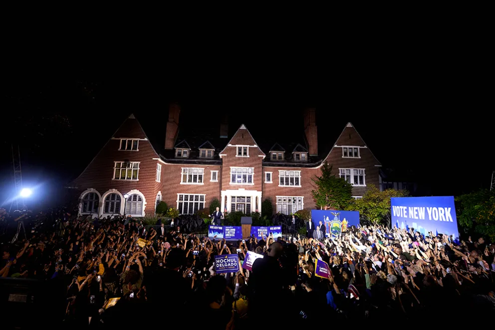 Large crowd gathered in front of a lit brick mansion at night, holding signs that read "Vote New York" and "Hochul Delgado" at a campaign rally.