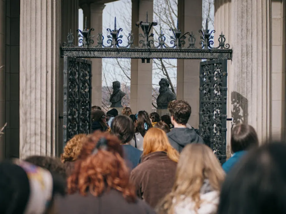 a group of people standing in front of a gate with statues of men and women on it and a crowd of people standing in front of the gate