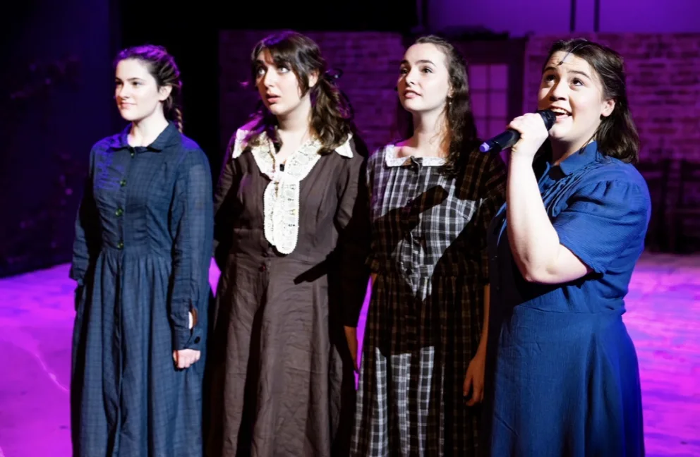 Four people in vintage-style dresses stand on stage, one holding a microphone, under purple and blue lights.