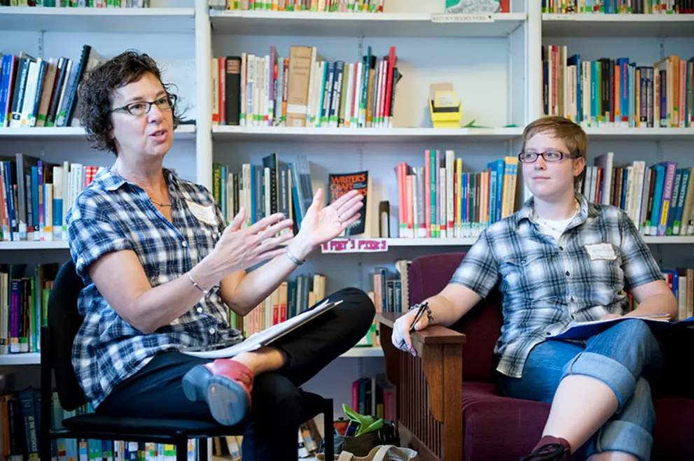 Two people sitting and talking in a library, both wearing plaid shirts, one gesturing with hands and the other holding a pen.