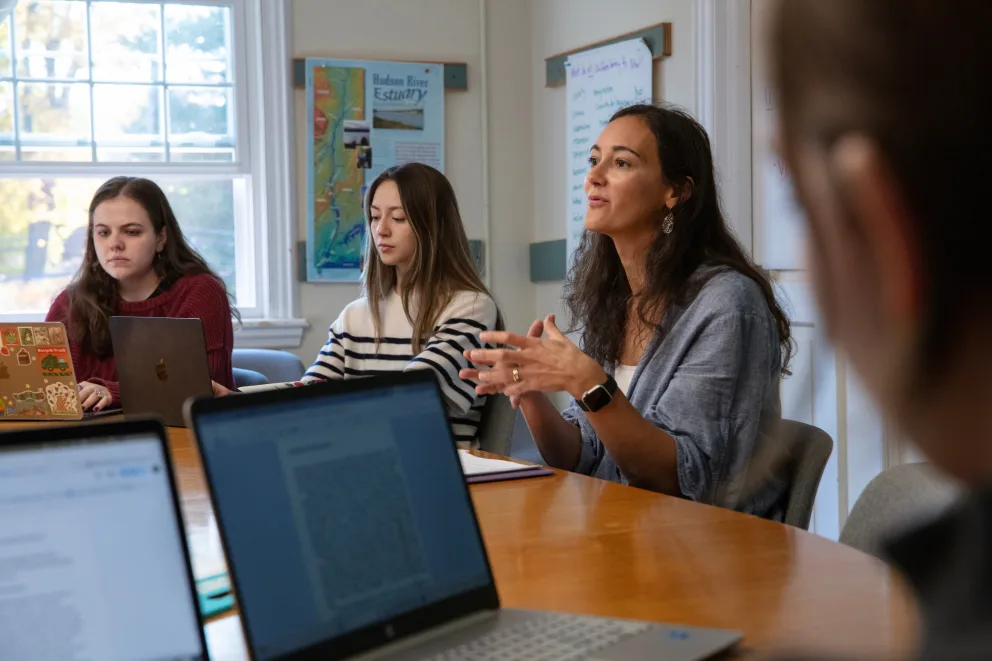 People sitting around a table with laptops, engaging in discussion. One person is speaking, while others listen attentively. Maps are on the wall.